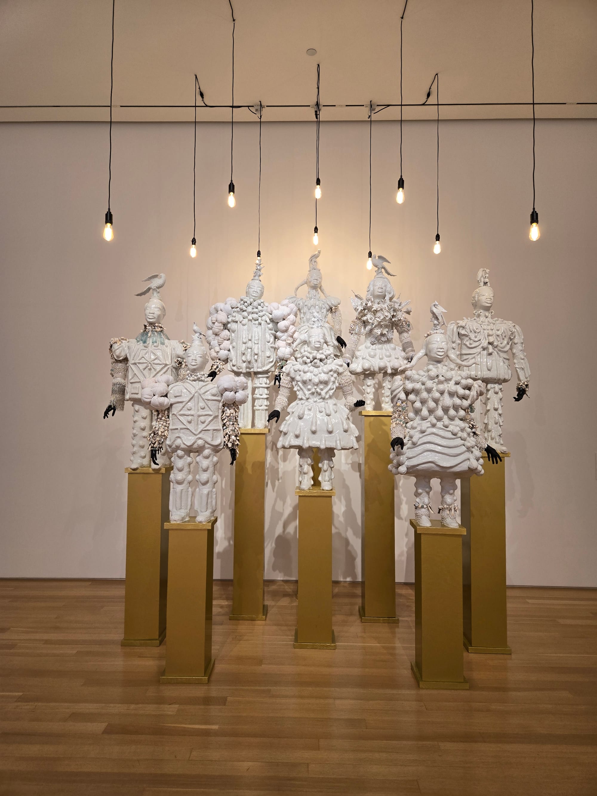 Eight sculptures entirely in white show small African-appearing people in elaborately decorated outfits, many with birds on their heads, and black gloves as hands stand on golden pedestals of differing heights. Bare light bulbs hang over the sculptures from the ceiling.