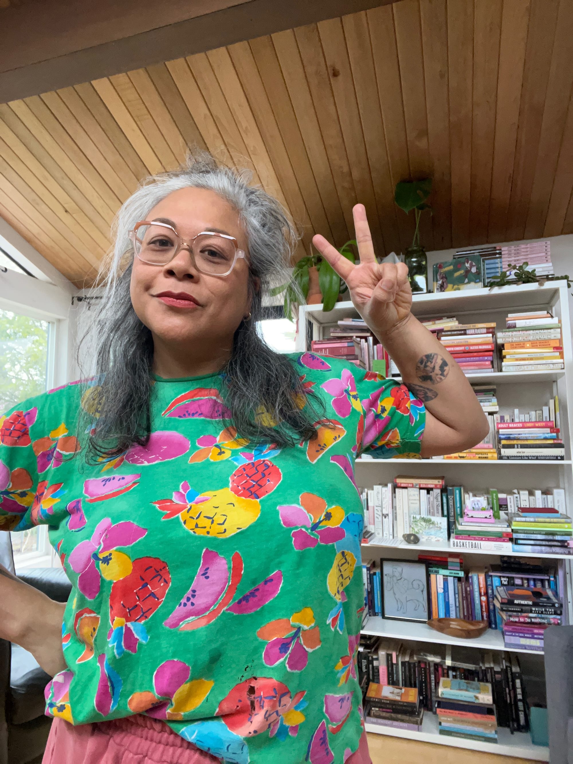 A filipina woman with gray hair and glasses holds up a peace sign while posing in a t shirt with a bright green background and bright painted fruit in front of a bookshelf.