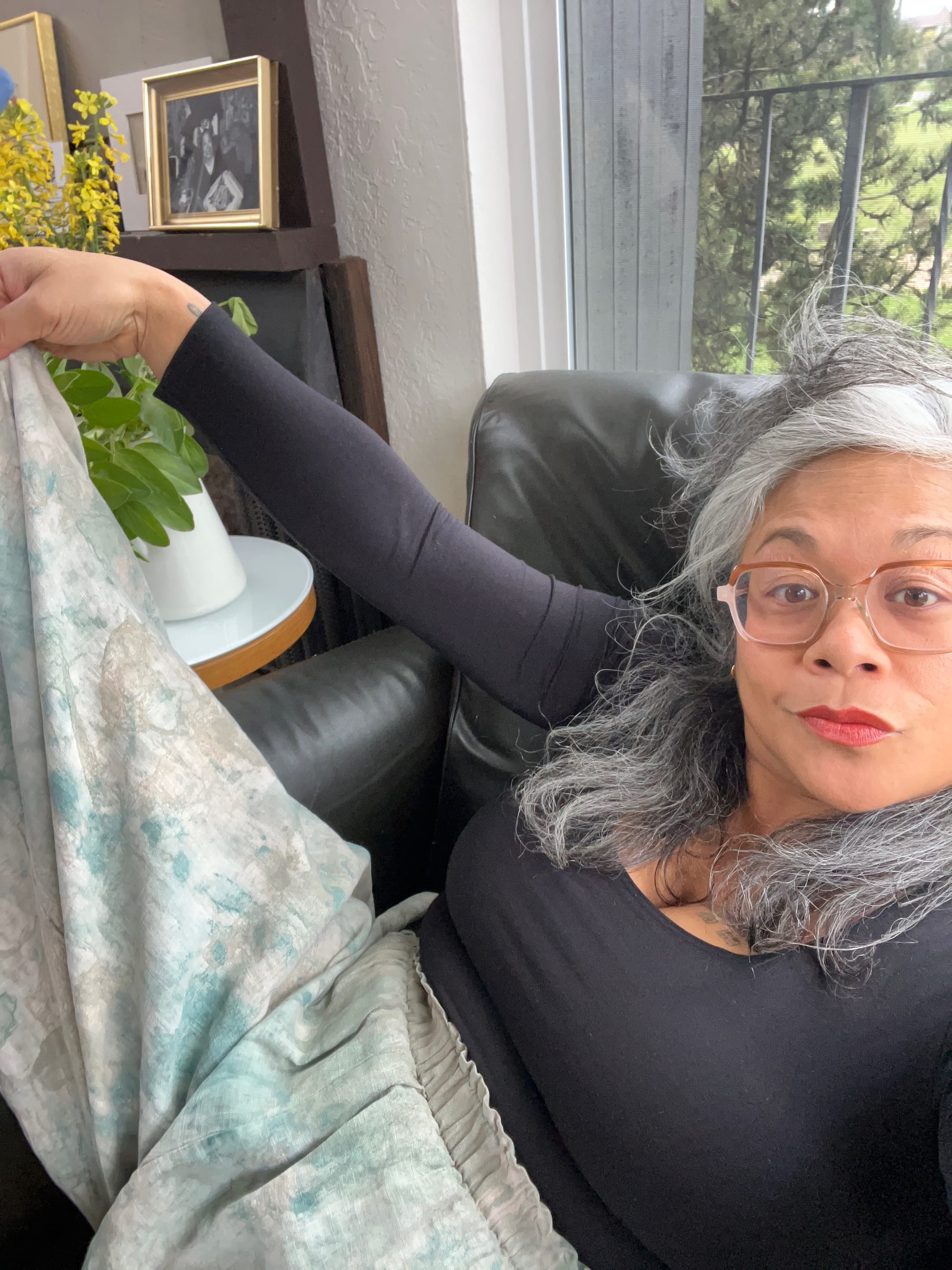 A brown skinned woman in glasses and a black shirt in a selfie holding up one leg of elastic waist hand painted pants.