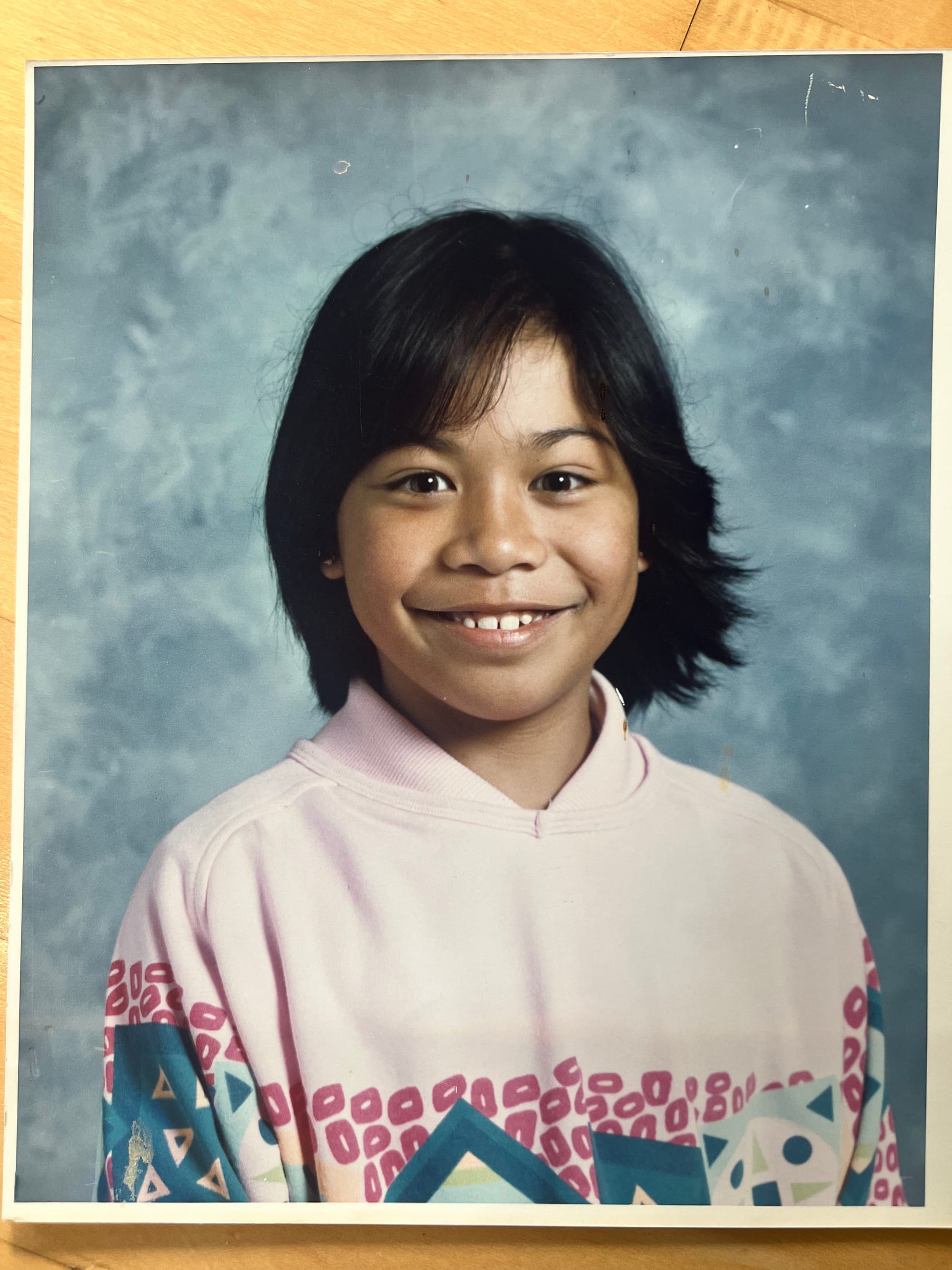 A young girl with brown skin and black hair cut in bangs and a short bob smiles in a school portrait while wearing a light pink sweatshirt with dark pink and turquoise shapes across the middle. 