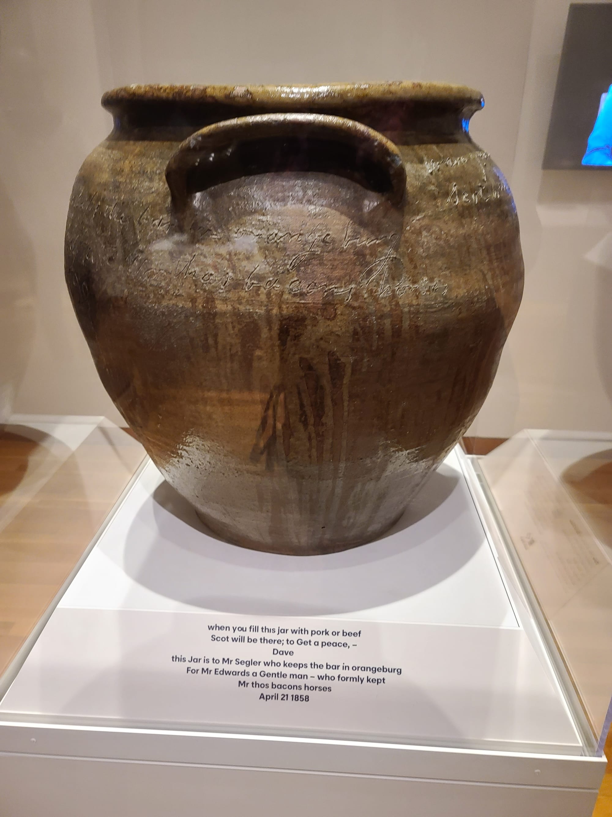 A brown clay storage jar with a handle is on a white display. There are words inscribed around the top edge of the pot.