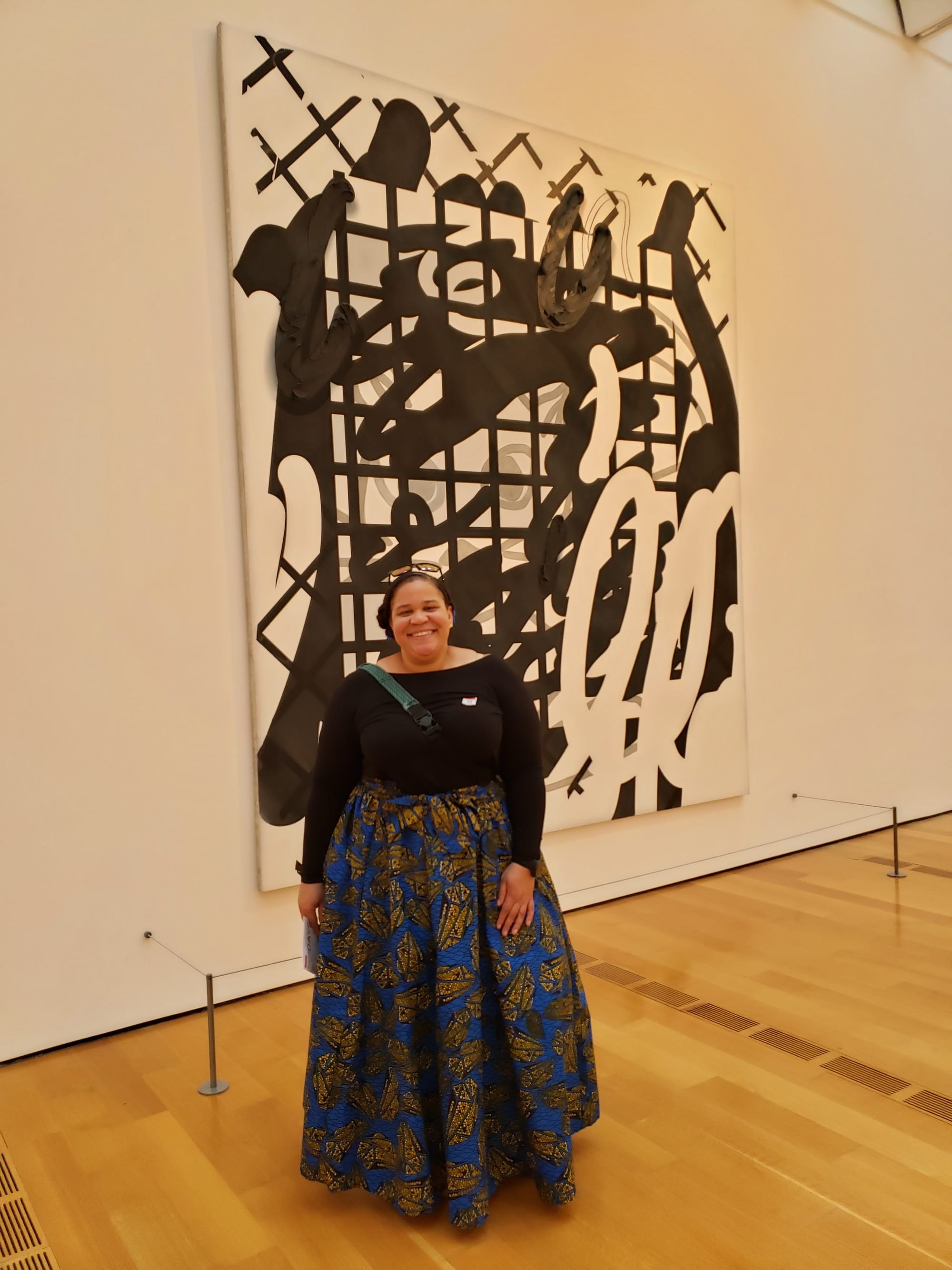 A smiling light-skinned Black woman wearing a black shirt and a blue and gold voluminous skirt stands in front of a black and white abstract painting on white walls.