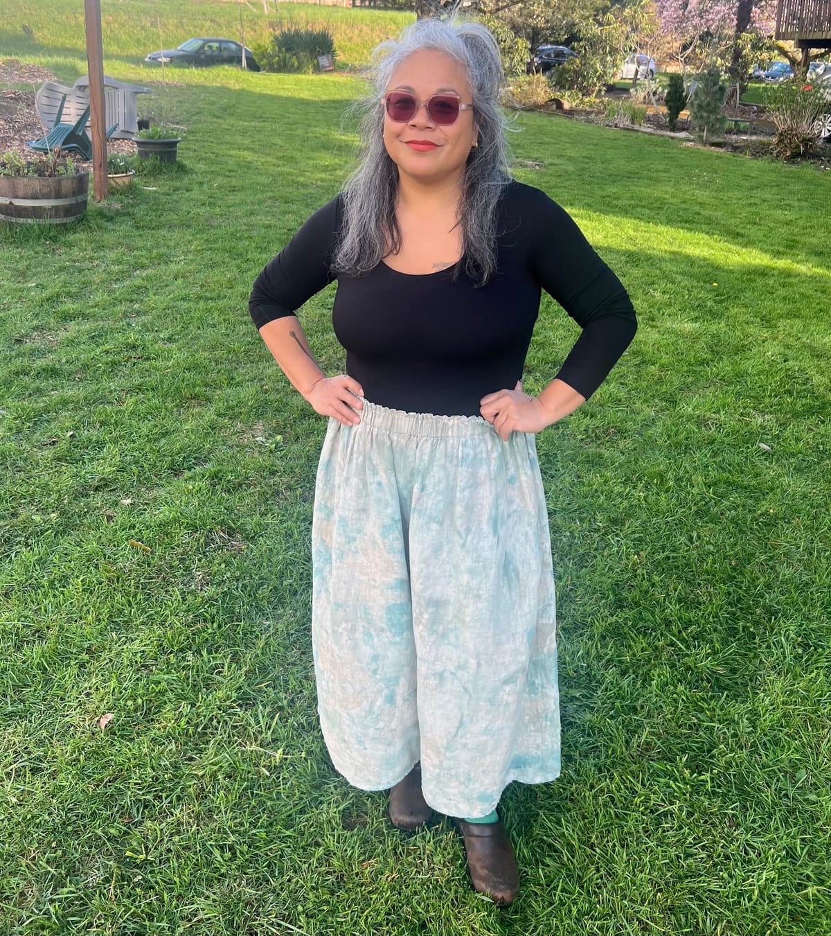 A brown-skinned woman with gray hair and sunglasses poses on a green hillside in teal painted linen pants and a black shirt with hands on hips.