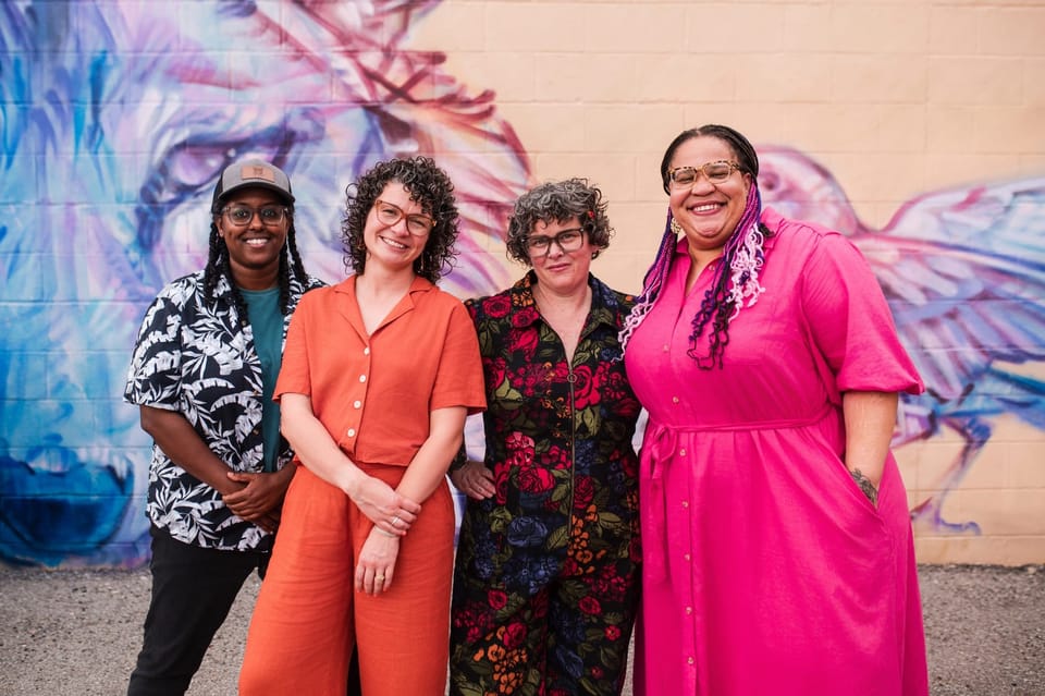 An outdoor group photo of four smiling people standing in front of a large mural. 